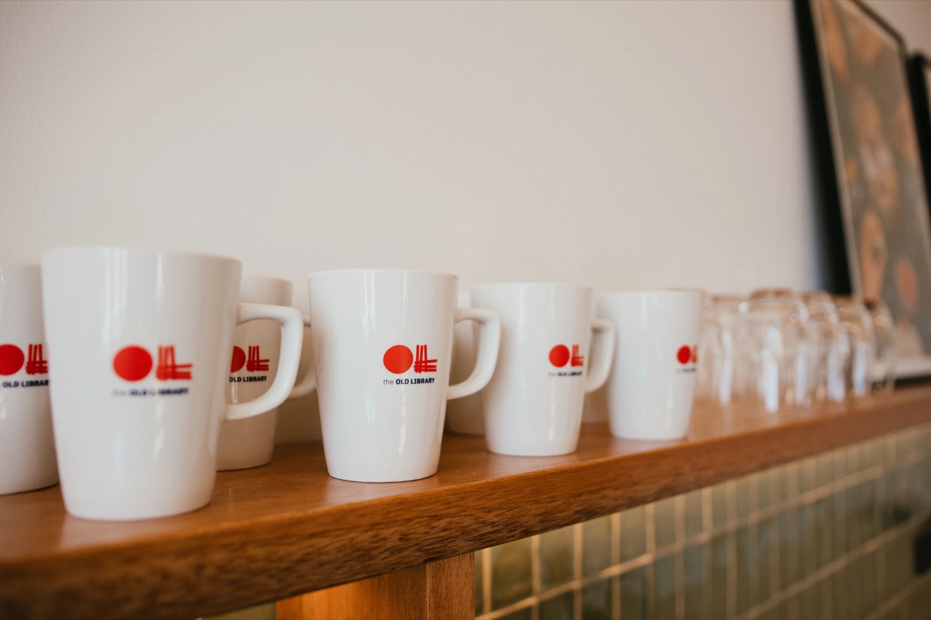 White mugs with The Old Library's logo lined up on a wooden shelf.