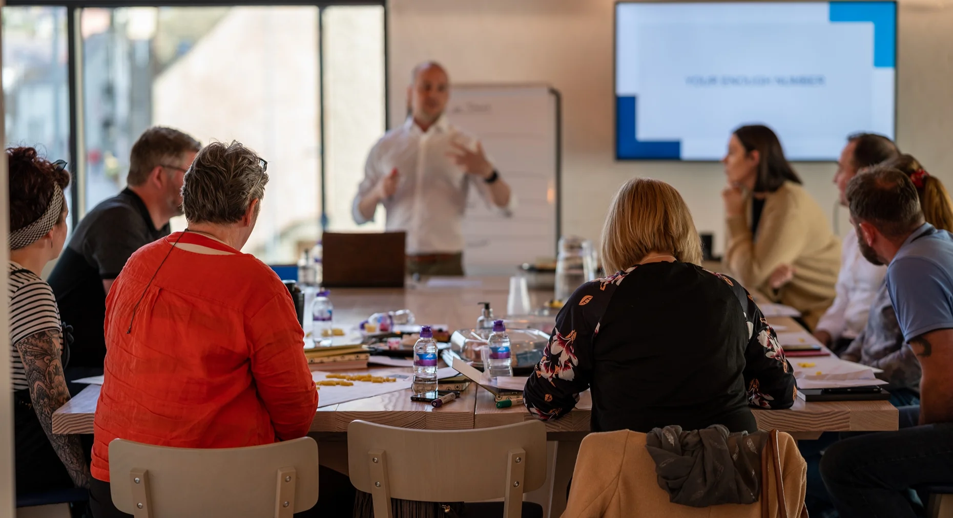 A workshop: people around a long table listening to a speaker in front of a screen.