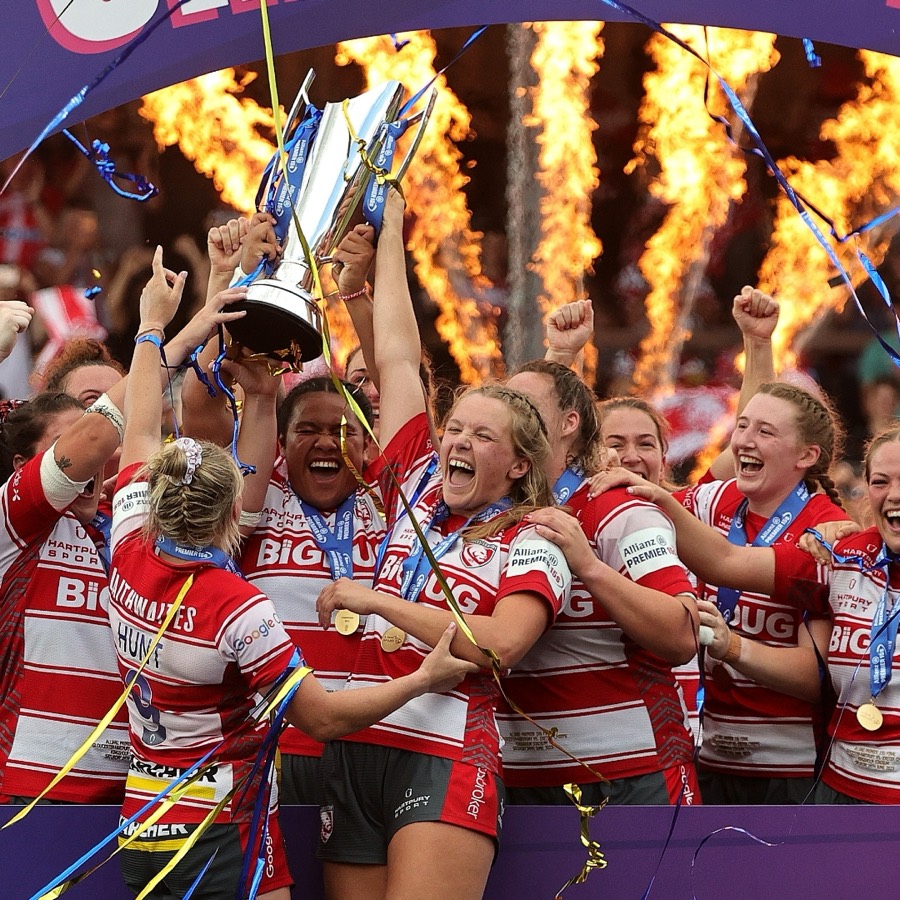 A women's rugby team lifts a trophy in front of flames and falling streamers.