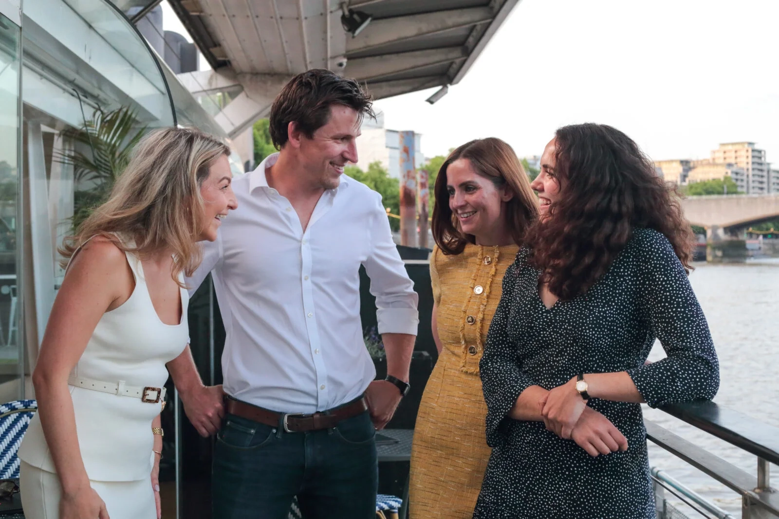 Four people laughing together on a riverside terrace.