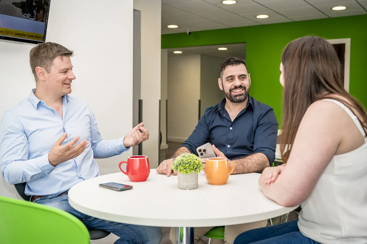 Chris talking with Ed and a colleague round a table in the office.