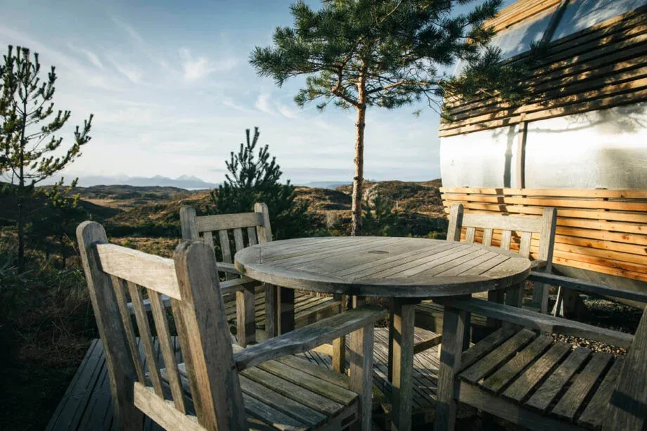 A wooden table and chairs on the cabin’s deck, with pines and distant hills.
