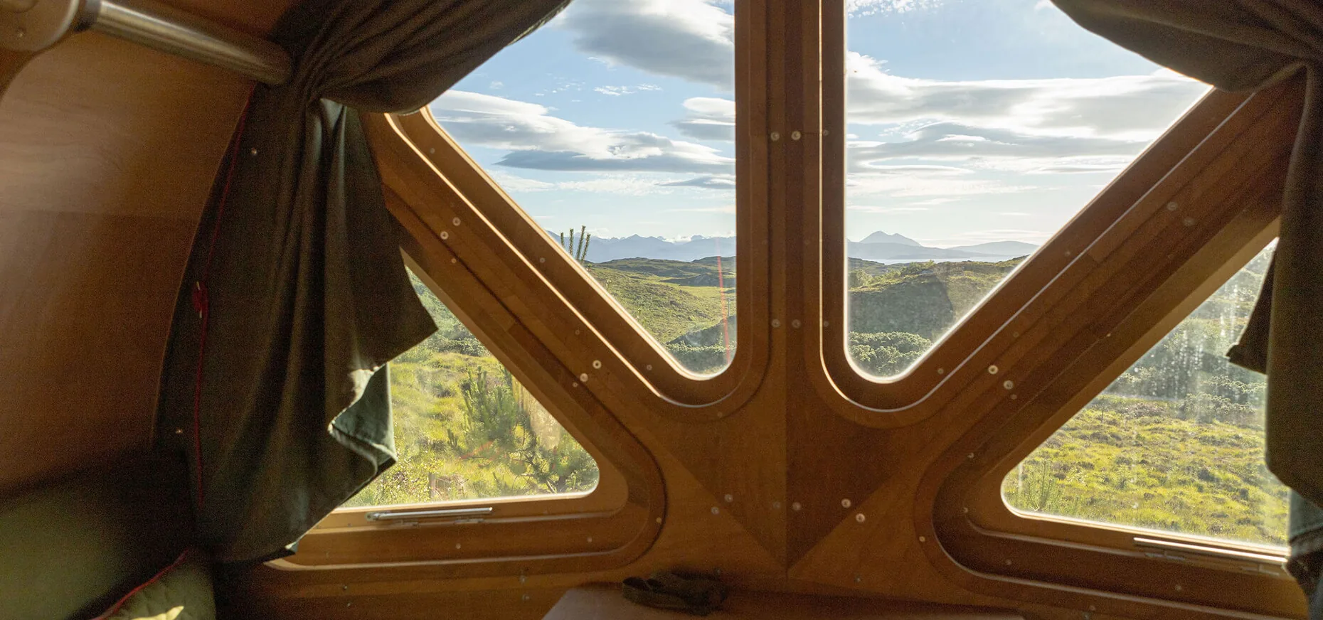 A view of green hills and sky through the cabin’s wooden-framed windows and curtains.