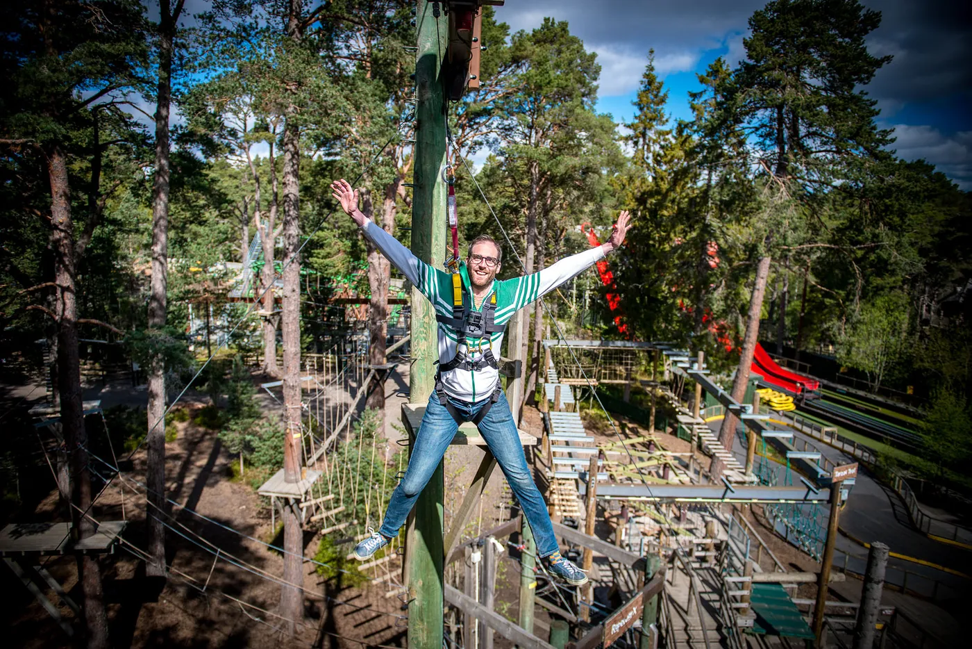 A man with arms spread wide jumping from a platform high in the forest, with climbing frames and a slide behind.