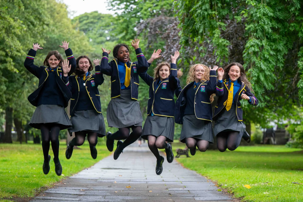 Six pupils in blazers jumping for joy together on a tree-lined path.